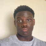 AJ wearing a grey crewneck shirt, posing against a neutral-colored wall.