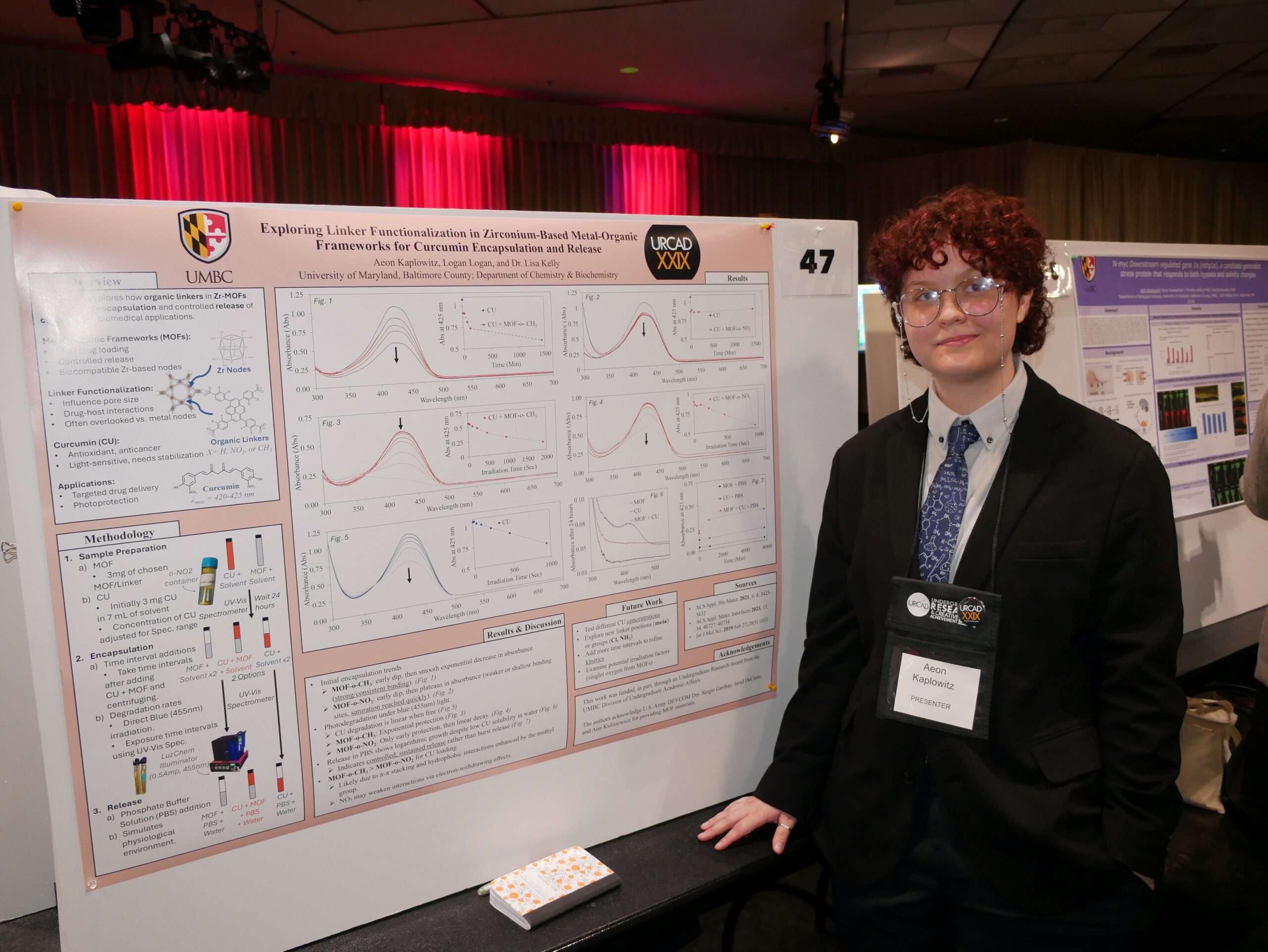 Aeon standing and standing next to URCAD poster project, in black blazer and blue tie with URCAD badge around neck