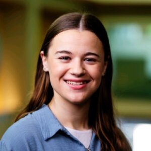 Miriam smiling and standing in front of a blue-green background.