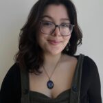 Amanda Negrete, smiling. She is wearing a dark green corduroy overalls and a circular blue pendant necklace against a white background.